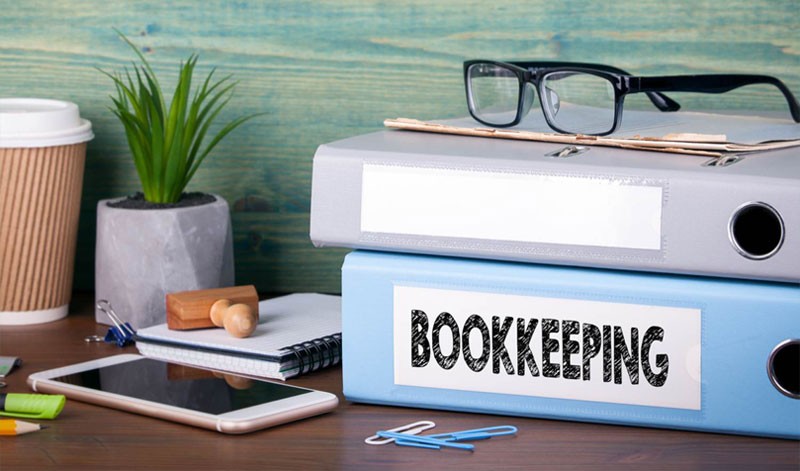 two books stacked on a desk with various items the bottom book is labeled bookkeeping